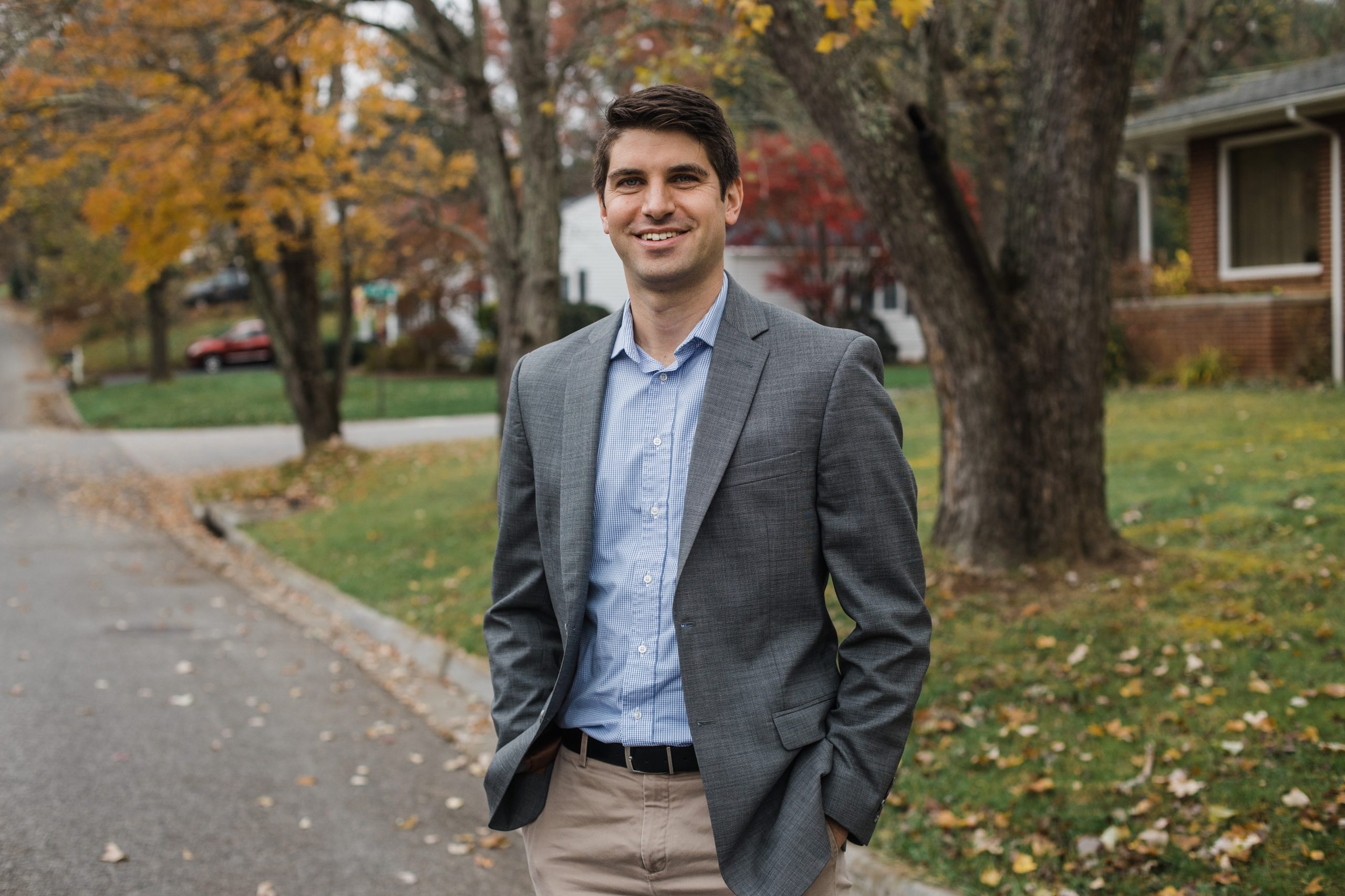 A man wearing a blazer smiles at a camera on a residential street in the Fall.